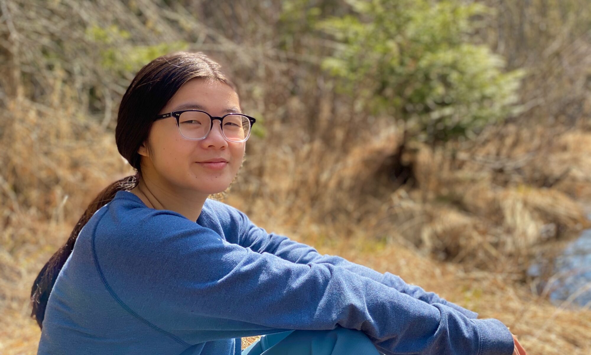 Headshot of Alyssa, a person with brown skin looking at the camera with a small grin, and sitting on the ground sideways with her knees bent, surrounded by a forested area. Alyssa has straight brown hair pulled back in a long ponytail, and she is wearing glasses and a blue long-sleeved top and pants.